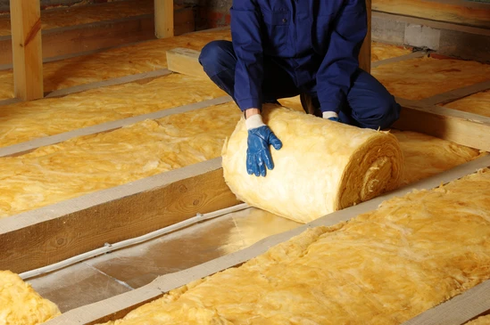 Ouvrier en combinaison bleue installant isolation thermique dans combles, manipulant rouleau de laine minérale jaune entre solives bois