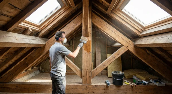 Photographie professionnelle montrant un artisan qualifié dans les combles ou sous la toiture d'une maison, traitant les éléments structurels de la charpente (poutres, solives) avec un produit de protection/traitement anti-xylophages. L'artisan porte des équipements de protection appropriés (masque, gants), utilise un équipement de traitement professionnel (pulvérisateur ou pinceau de traitement spécialisé), et travaille sur les bois de structure visibles. La lumière naturelle provenant des ouvertures de toiture éclaire la scène et met en évidence le travail de traitement en cours.