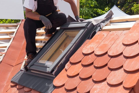 Ouvrier en salopette noire installant une fenêtre de toit sur une toiture en tuiles terre cuite rouge